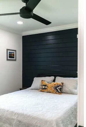 Une chambre à coucher avec un lit, un ventilateur de plafond, un tableau encadré accroché au mur et une tête de lit sombre.