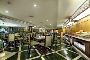 Comedor de hotel con varias mesas y sillas, una barra de buffet con diversos alimentos, y un mostrador de recepción al fondo.