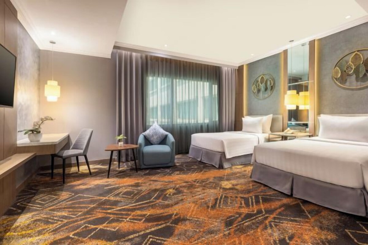 Habitación de hotel con cama, dos sillas, escritorio, televisor y una ventana grande con cortinas.