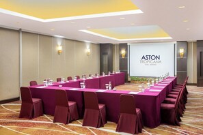 Sala de conferência com uma mesa comprida, cadeiras e uma tela exibindo o nome do hotel.