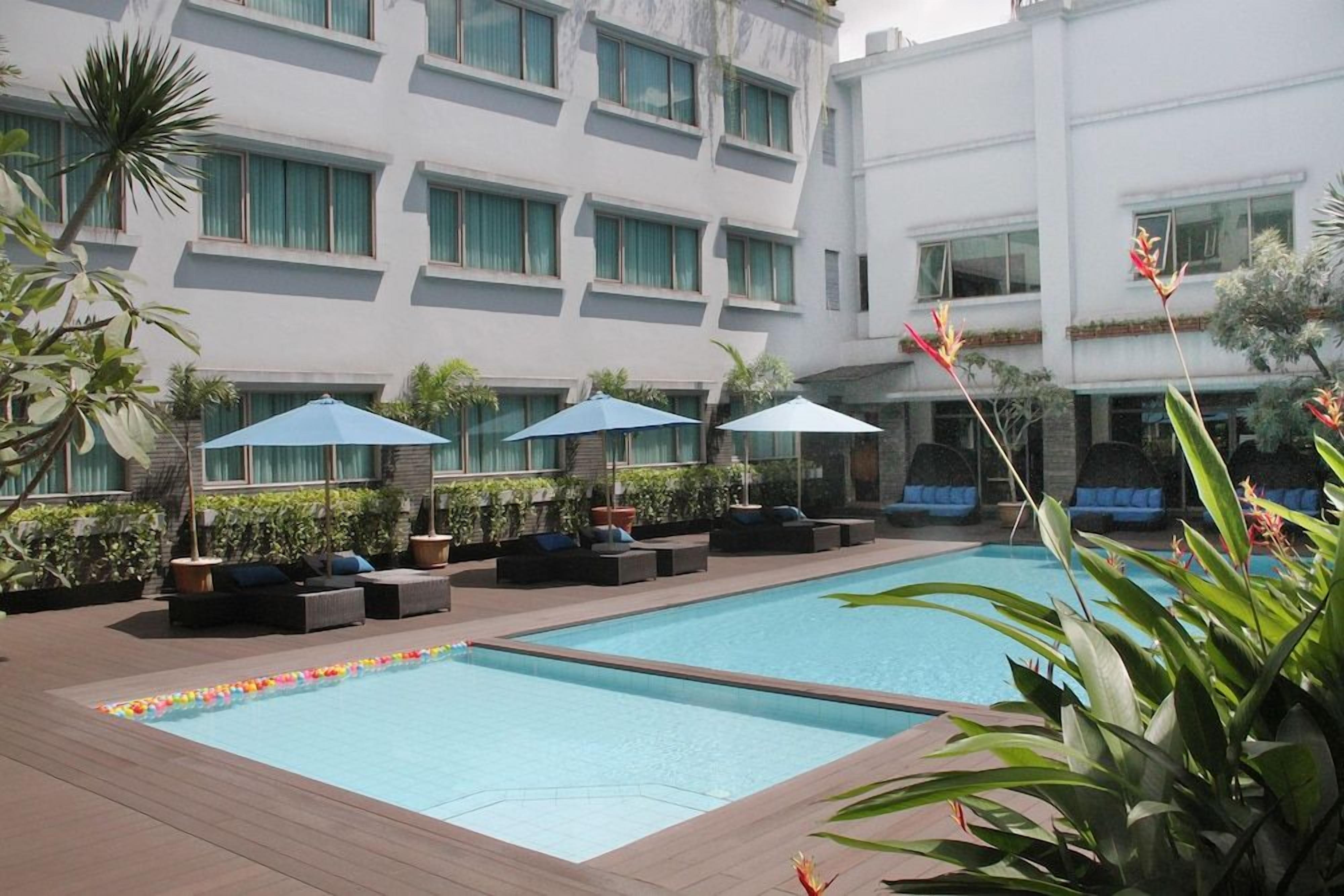 A swimming pool with lounge chairs and umbrellas, surrounded by a building with multiple floors and windows.