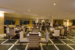 Ein Hotel-Speisebereich mit mehreren Tischen und Stühlen, einem zentralen Buffet und einem gemusterten Boden.