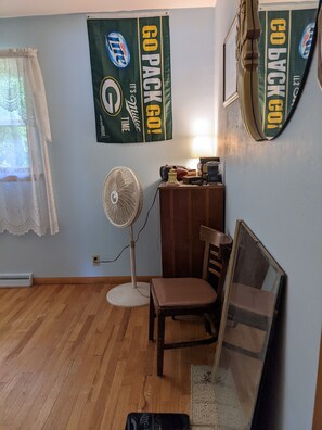 Une pièce avec un plancher en bois, un bureau avec une lampe, une chaise, un ventilateur et deux affiches au mur.