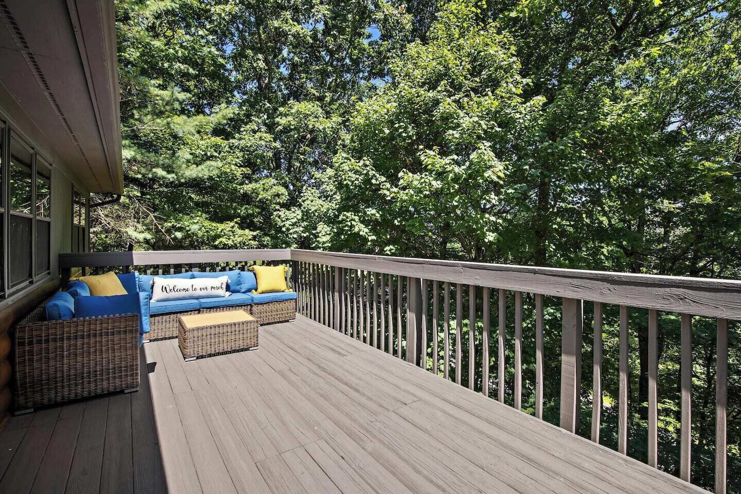 Une terrasse en bois avec un canapé bleu et des coussins jaunes, entourée d’arbres.