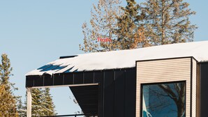 A modern building with a sloped roof covered in snow, wooden exterior panels, and a large window reflecting trees.