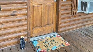 Una cabaña de madera con una puerta grande, un pequeño equipo de aire acondicionado y un felpudo colorido.