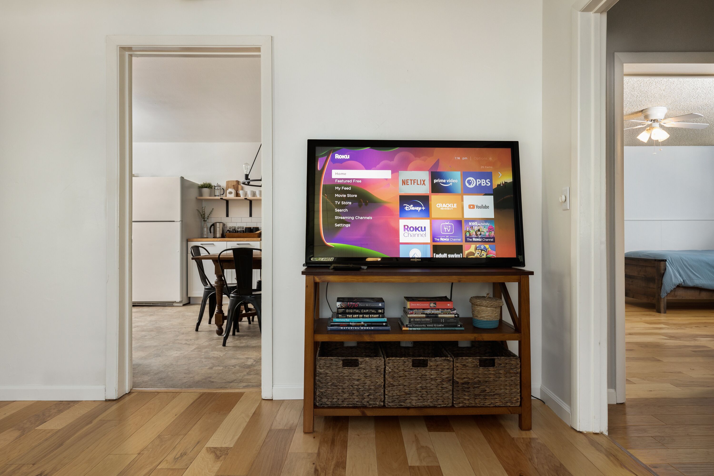 A living room with a flat-screen TV on a wooden stand, a dining area with a table and chairs, and a bedroom with a bed.