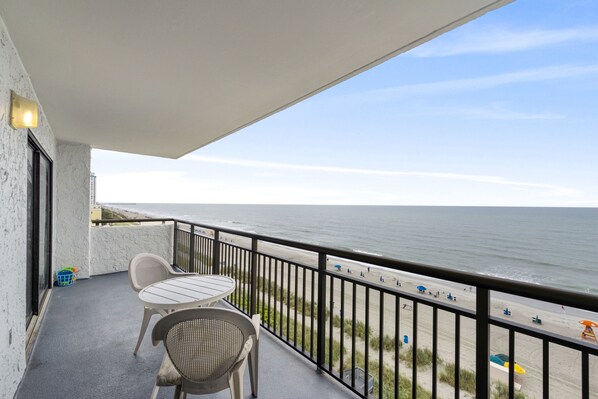 A balcony with a table and chairs, overlooking a beach with umbrellas and people. - Amazing Large Oceanfront Sleeps (8) -2 bedroom 2bath (Myrtle Beach)