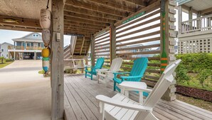 A covered outdoor seating area with white and turquoise Adirondack chairs, a wooden deck, and a view of a residential building in the background. - Bright and sunny family friendly cottage, 2 minutes to beach, Sound view sunsets (Kill Devil Hills)