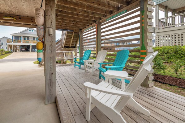 A covered outdoor seating area with white and turquoise Adirondack chairs, a wooden deck, and a view of a residential building in the background.