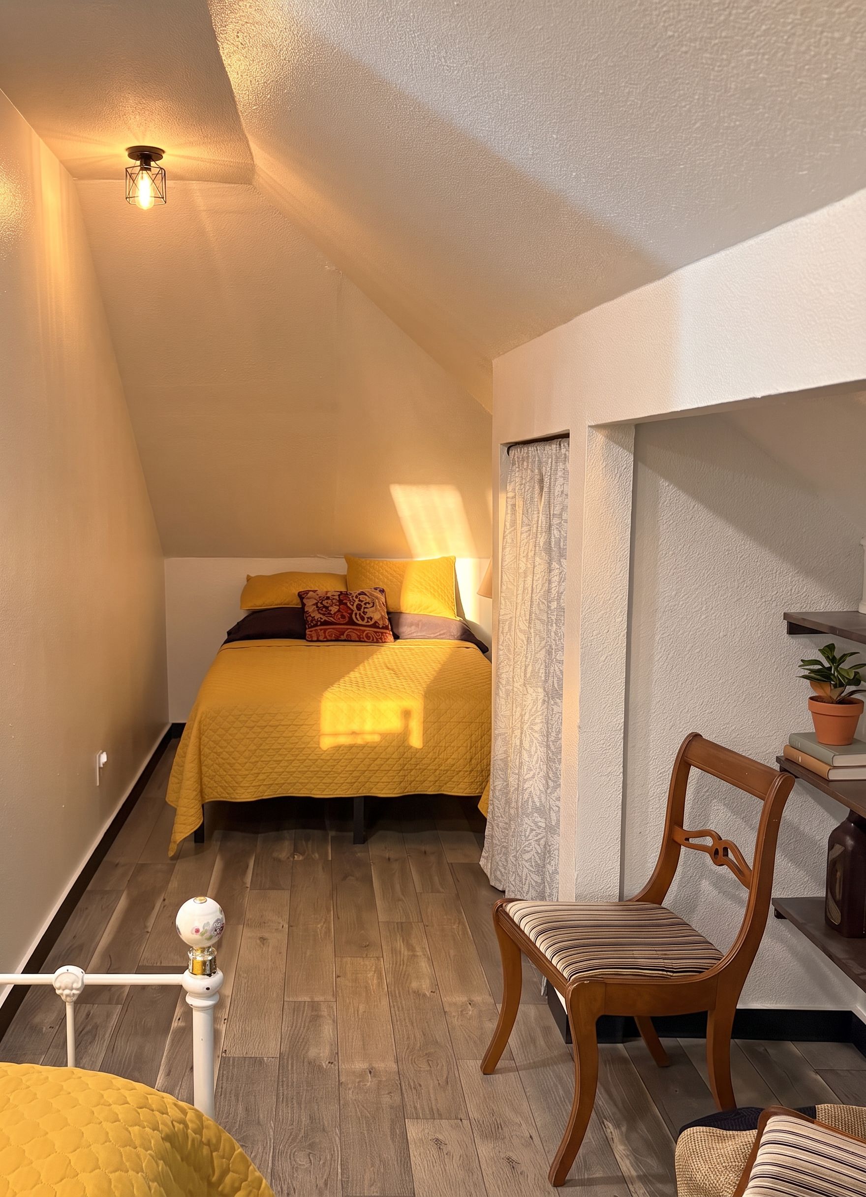 A bedroom with a bed, a chair, a small table with a plant, and a window with curtains.