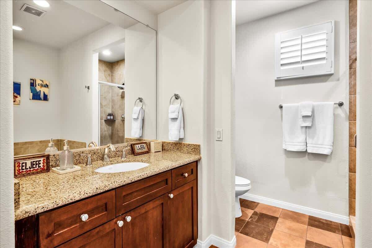 A bathroom with a granite countertop sink, wooden vanity, and a large mirror. There are two towels hanging on the wall and a shower area visible in the mirror reflection.
