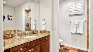 A bathroom with a granite countertop sink, wooden vanity, and a large mirror. There are two towels hanging on the wall and a shower area visible in the mirror reflection.