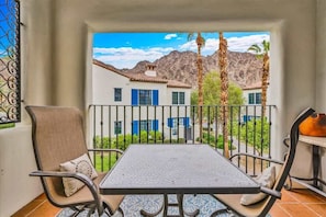 A balcony with a table and chairs, a view of a mountain, and a building with blue shutters. - Desert Sky Vista B | Studio Legacy Villa (La Quinta)