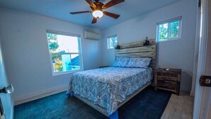 Un dormitorio con cama, ventilador de techo, dos ventanas y mesita de noche.