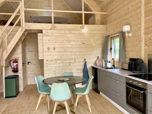 Ein gemütliches Blockhaus-Interieur mit Holzvertäfelung, einem Essbereich mit rundem Tisch und Stühlen, einer Küche mit Herd und Spüle sowie einer Treppe zu einem Schlafbereich auf dem Lofts.