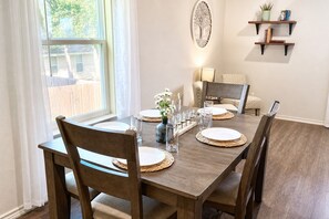 A dining area with a wooden table set for four, surrounded by chairs, with a window and a wall-mounted shelf with books and a plant.