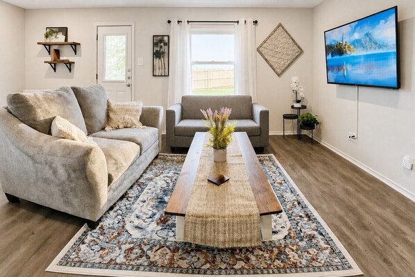 A living room with a sofa, armchair, coffee table, and a TV on the wall.