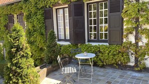 Un patio couvert, agrémenté d’une table et de chaises, entouré de verdure et bordé d’un bâtiment doté de fenêtres et de volets.