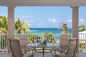 Un espace extérieur abrité, doté d’une table ronde et de quatre chaises, offrant une vue sur une plage bordée de palmiers et sur l’océan.