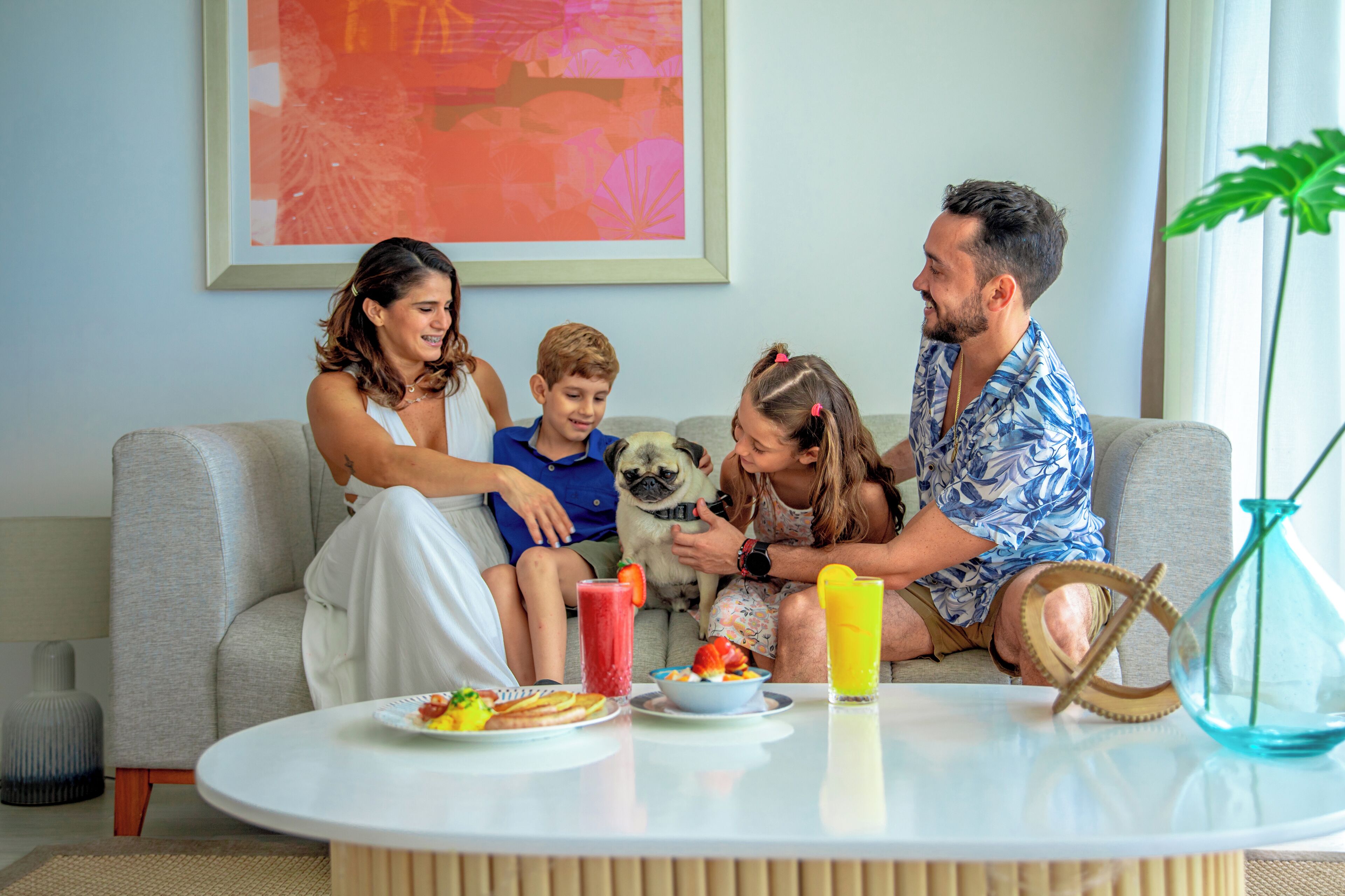 A family sitting on a grey sofa with a table in front of them holding food and drinks.