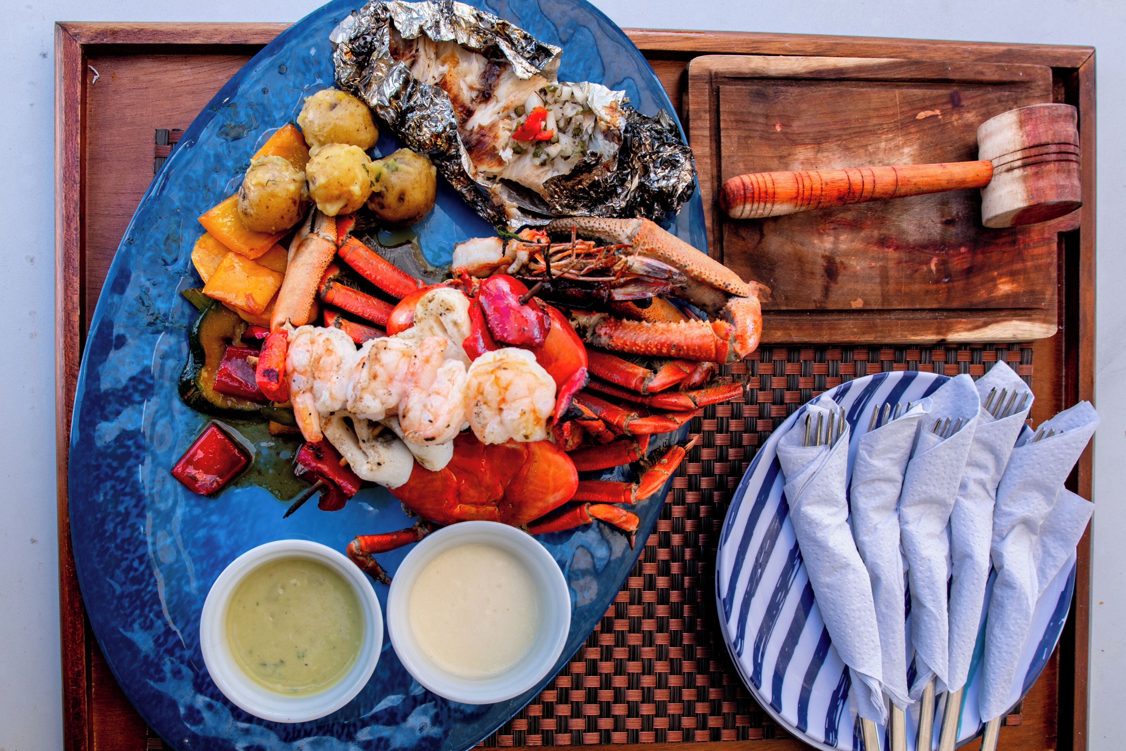 A seafood platter with various grilled items, including shrimp, crab, and vegetables, accompanied by dipping sauces and a wooden mallet.