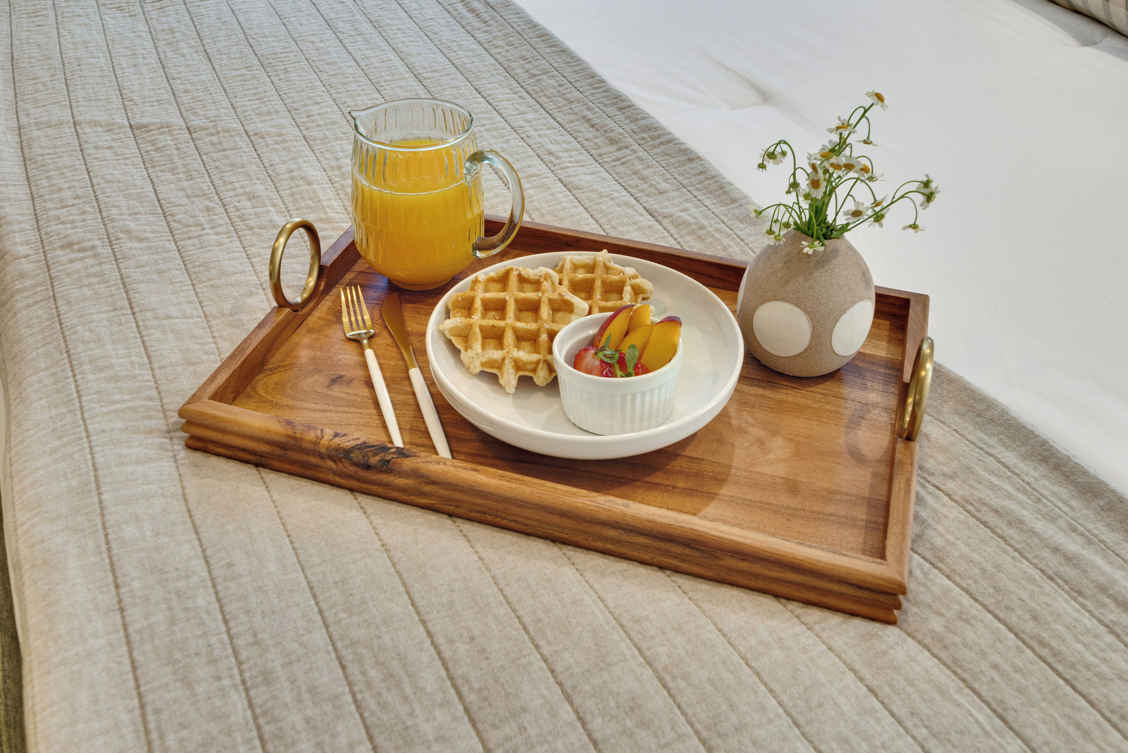 A wooden tray with waffles, a glass of orange juice, a bowl of fruit, and a vase with flowers.