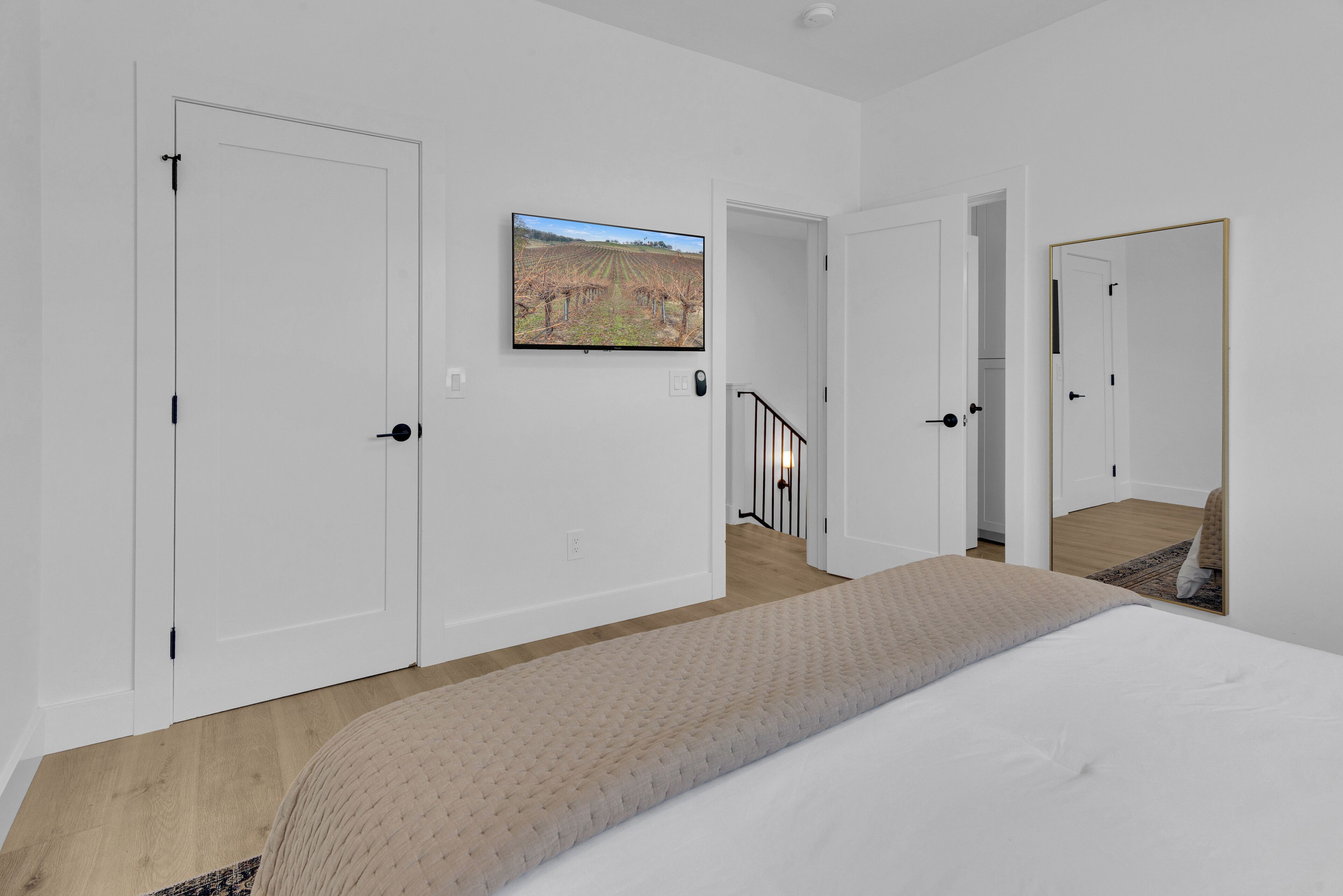A bedroom with a bed, a TV mounted on the wall, and a view of a vineyard through an open door.