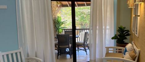 A room with a sliding glass door leading to an outdoor patio, two white chairs, a small table, and a potted plant.