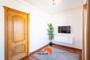 Una habitación con puerta de madera, planta en maceta, televisor montado y alfombra gris.