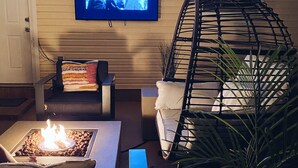 Ein gemütliches Wohnzimmer mit flachbildfernseher an der Wand, einer bequemen Couch, einem kleinen Tisch mit Feuerstelle und einem Hängekorbstuhl.