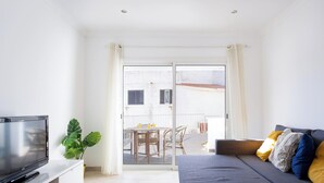 Una sala de estar moderna con un televisor de pantalla plana, un sofá gris con cojines amarillos y azules, una planta verde grande y un ventanal corredizo que da a un patio exterior con una mesa y sillas.