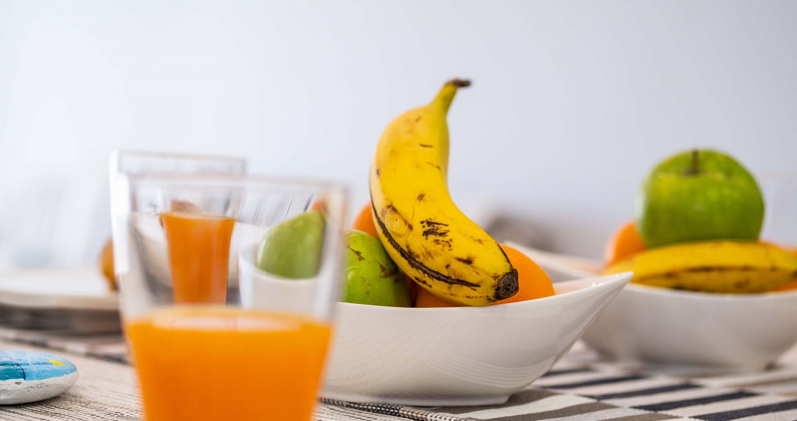 A breakfast setting with a glass of orange juice, a bowl of fruits including bananas and apples, and a cup of coffee.