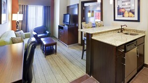 Habitación de hotel con comedor, sofá, televisor y cocina con fregadero, además de un cuadro en la pared.