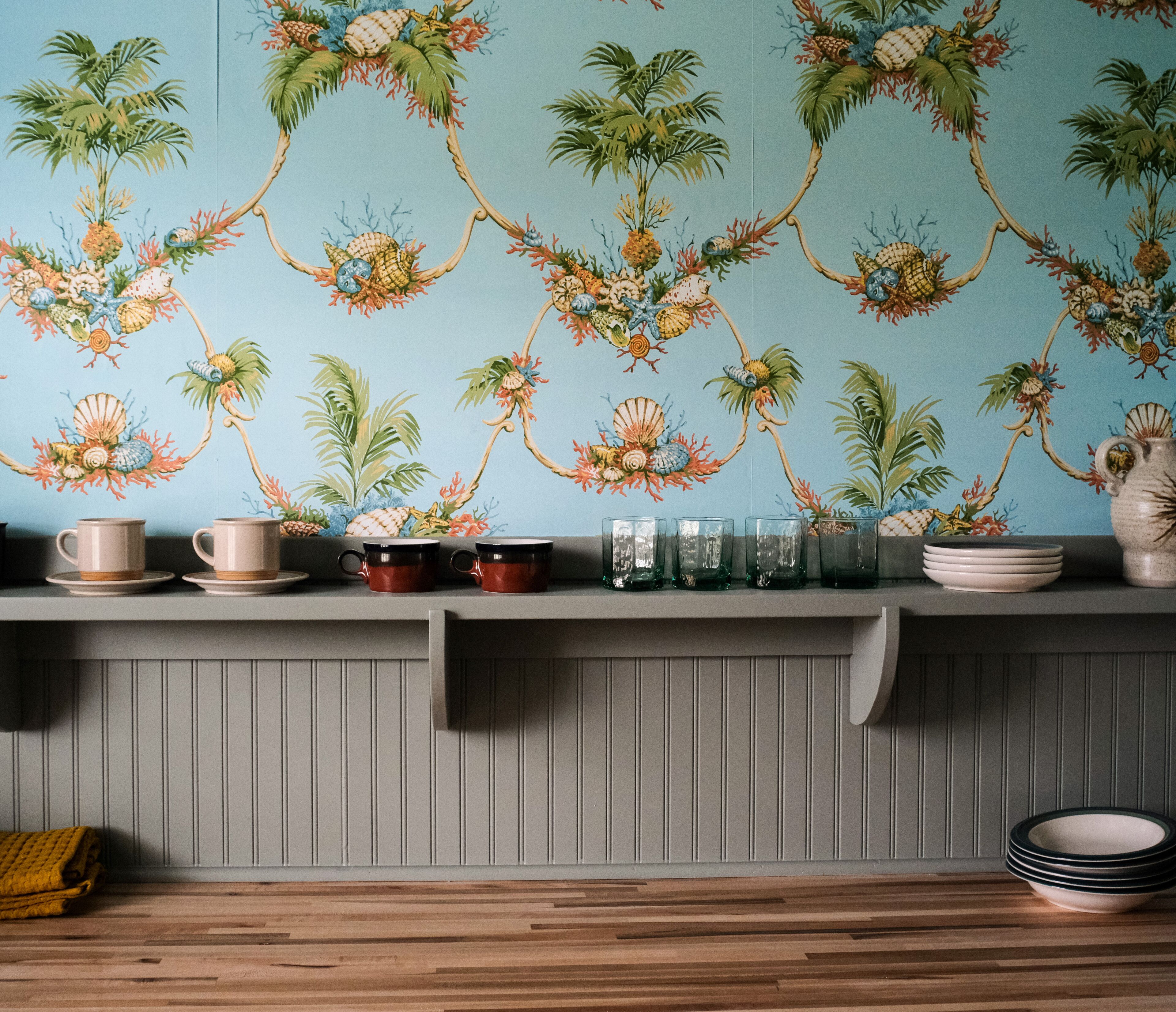 A kitchen with a blue floral wallpaper, wooden shelves with various dishes, and a wooden floor.