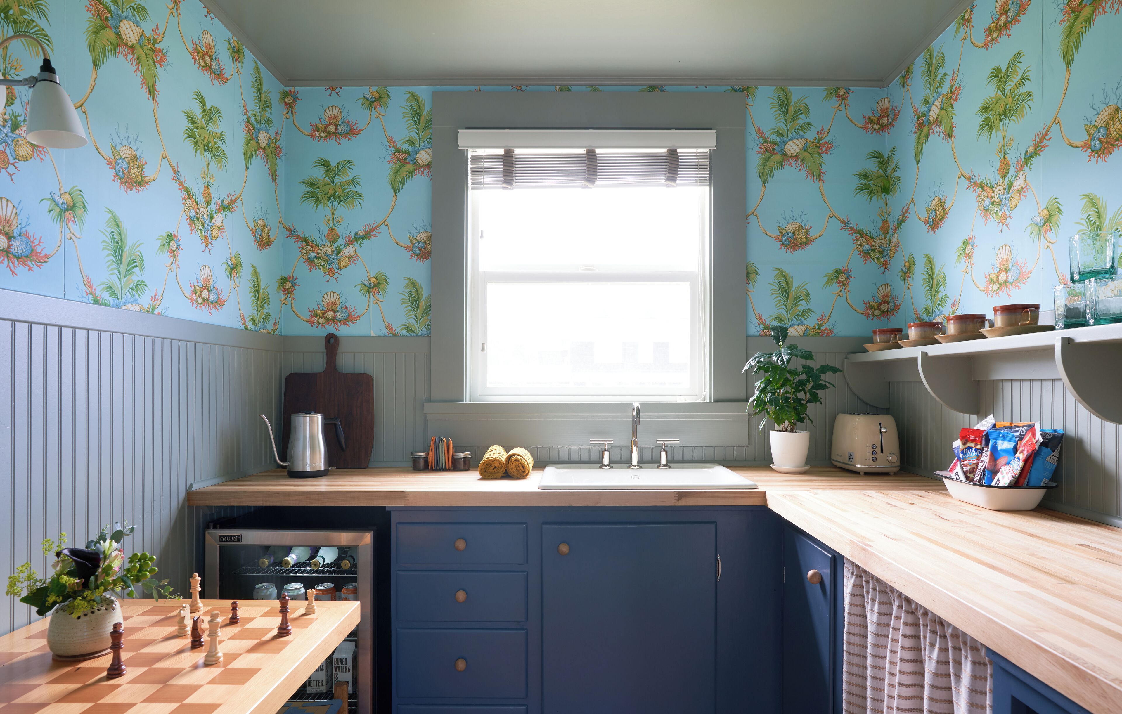A kitchen with a wooden countertop, a sink, and a window with blinds. There is a chessboard on the counter and a plant in a pot.