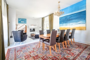 Un coin repas avec une table et des chaises en bois, un tapis à motifs et un grand tableau représentant une scène de plage.