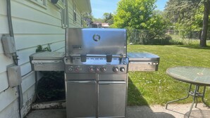 Un barbecue à gaz Weber Summit en acier inoxydable, doté de quatre brûleurs, d’une étagère latérale et d’un thermomètre intégré, placé sur une terrasse en béton.