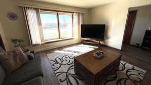 Un salon comprenant un canapé, une table basse, un téléviseur à écran plat et une grande fenêtre avec des rideaux.