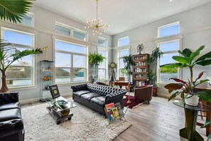 Relax in this spacious living room with a high ceiling and large windows, featuring a comfortable black leather sofa, glass coffee table, and bookshelves filled with books and decorative items.
