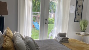 Ett rum med en säng, ett litet bord med en växt och en skjutdörr i glas som leder ut till en trädgård med en rutschkana och grönska.