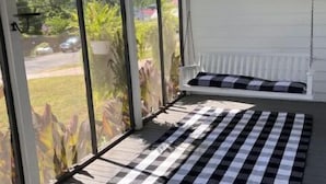 A covered porch with a checkered mat, a white bench with cushions, and a view of greenery and a street. - The Jacks Fork Town House in Eminence (Eminence)