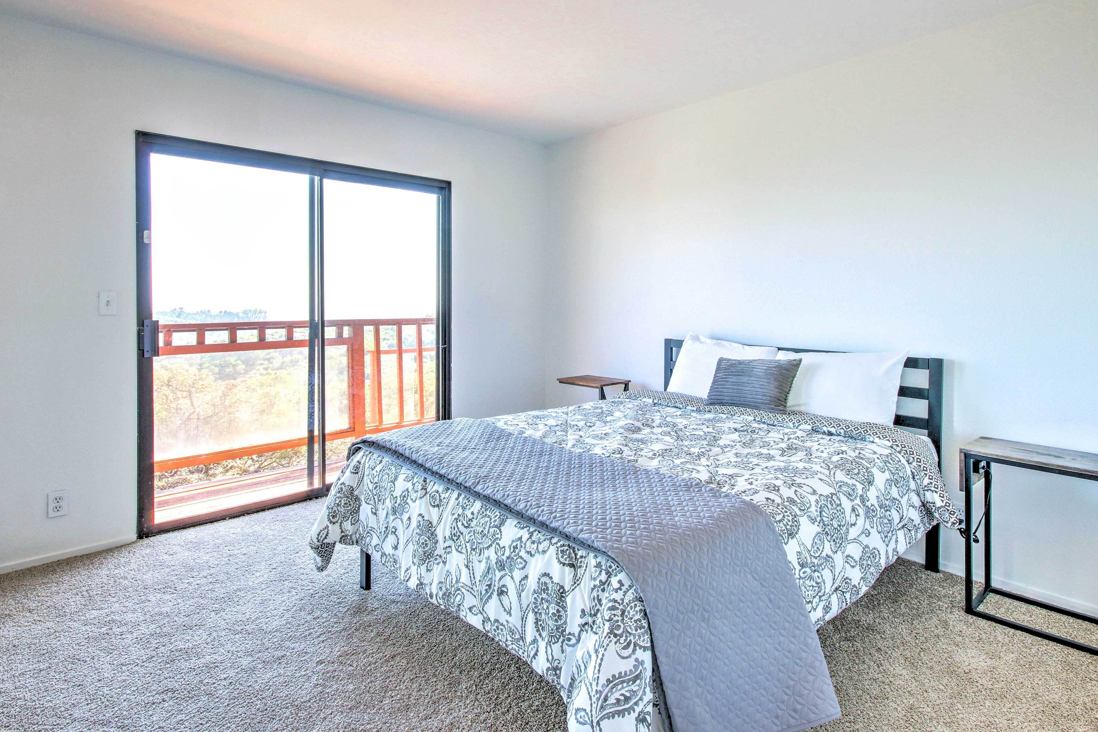 A bedroom with a large bed, a nightstand, a window with a view, and a sliding door to a balcony.