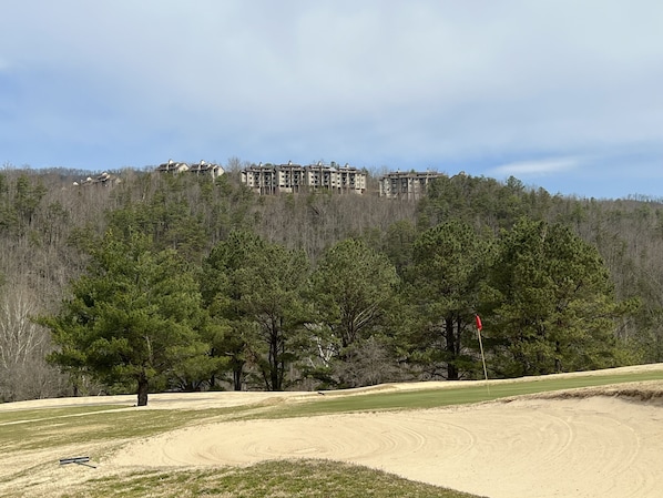 A golf course with sand bunkers, a flagstick, and a forested hill with buildings in the background.