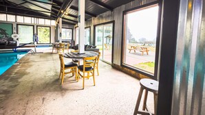 A covered outdoor area with a pool, wooden tables and chairs, and large windows offering views of the outside. - Exclusive Cottage & Coastal Farmhouse Private Indoor Heated Pool ocean views. (Smith River)