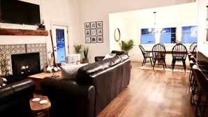 A living room with a fireplace, a black leather sofa, a dining area with a table and chairs, and a kitchen in the background.
