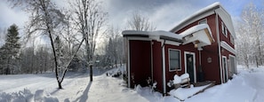 A red cabin with a snow-covered roof, surrounded by trees and a snow-covered ground.