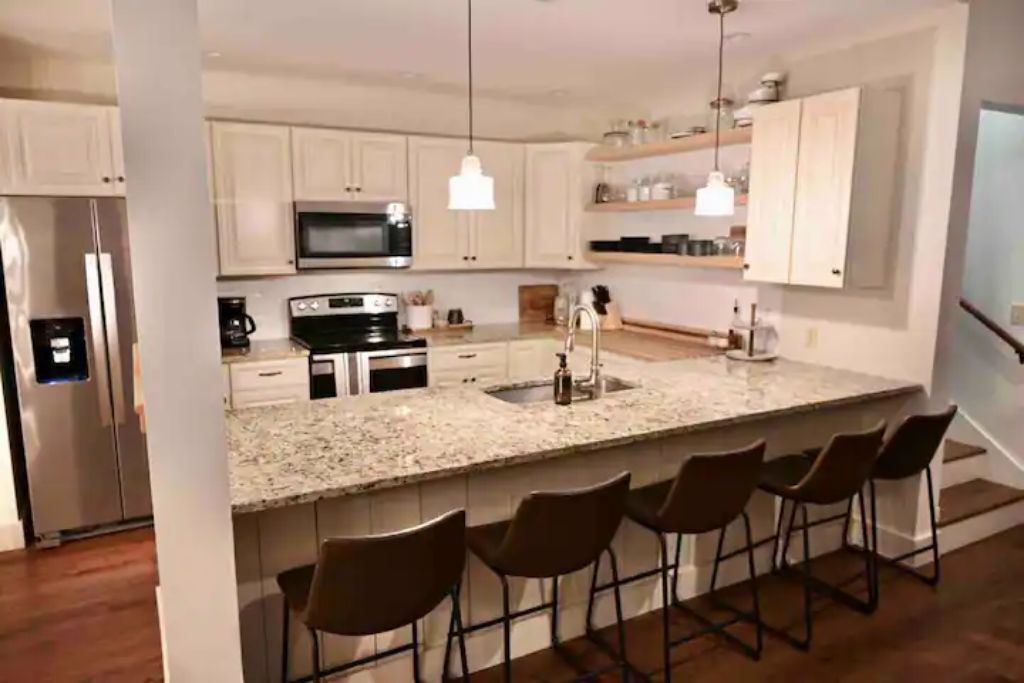 Una cocina moderna con armarios blancos, encimera de granito, microondas integrado, refrigerador de acero inoxidable y una barra con cuatro taburetes.
