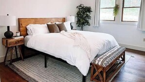 A bedroom with a wooden headboard, white bedding, a bedside table with a lamp, a bench, and a large window with curtains.