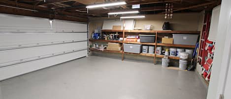 A garage with concrete floors, wooden beams, fluorescent lighting, and shelves with storage bins and tools.